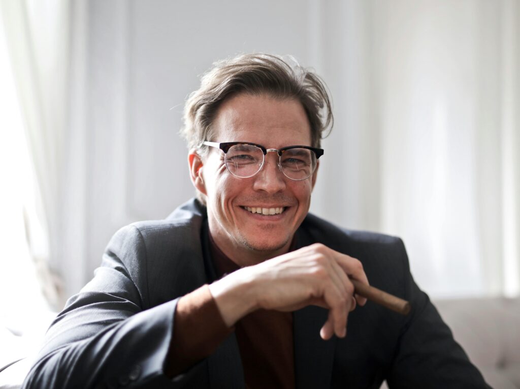 Cheerful businessman in glasses and suit holding a cigar, indoors.
